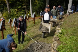 Kilku mężczyzn i kobiet pracuje na stromym cmentarzu wojennym, wysypując kamień z taczek i rozgarniając go grabiami między rzędami krzyży. To zbiorowa akcja porządkowa prowadzona na pochyłym terenie pod lasem.