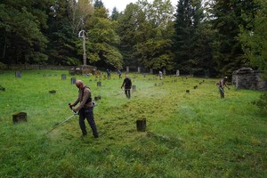 Na dużej, zielonej polanie cmentarnej kilku mężczyzn kosi wysoką trawę podkaszarkami między kamiennymi nagrobkami. W tle stoi wysoki krzyż i otaczający cmentarz drewniany płot.