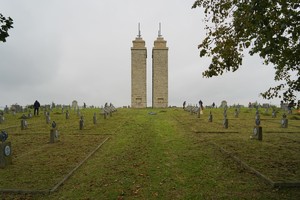 Szeroki kadr na znany cmentarz wojenny z dwiema wysokimi kamiennymi wieżami stojącymi na wprost wejścia. Na uporządkowanym terenie między grobami pracuje kilka osób, co podkreśla skalę nekropolii.