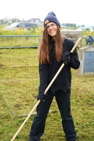 Młoda kobieta w ciemnym stroju, czapce i rękawicach stoi z grabiami na uporządkowanym cmentarzu. Za nią widać kamienne nagrobki i metalowe ogrodzenie, a w tle samochody i zabudowania.