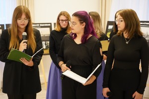Grupa młodych kobiet w czarnych strojach występuje z mikrofonami i śpiewnikami podczas części artystycznej spotkania. W tle stoją plansze z fotografiami cmentarzy wojennych.