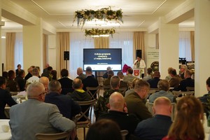 Widok ogólny sali konferencyjnej, gdzie uczestnicy siedzą przy stołach i słuchają wystąpienia na tle dużego ekranu. Na ekranie widać tytuł wydarzenia i rok, a obok stoją roll-upy organizatorów.