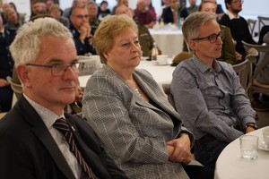 Zbliżenie na uczestników siedzących przy stolikach i słuchających wystąpień; widać kobietę w kraciastym żakiecie i dwóch mężczyzn po bokach. Tło jest nieostre, co podkreśla skupienie słuchaczy.