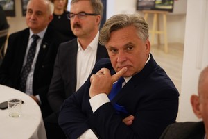 Mężczyzna w granatowym garniturze i z niebieskim krawatem siedzi przy stole i uważnie słucha prelegentów. W tle znajdują się pozostali goście spotkania w sali konferencyjnej.