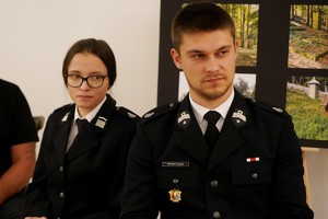 Na zdjęciu siedzą obok siebie dwie młode osoby w galowych mundurach, patrzące w stronę mówiącej osoby. W tle widać plansze z fotografiami cmentarzy wojennych.