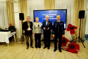 Cztery osoby stoją na tle ekranu z napisem „X Akcja sprzątania cmentarzy z I wojny światowej”, trzymając w dłoniach odznaczenia. To moment uhonorowania osób za pracę przy akcji.