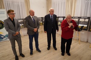 Czterech mężczyzn stoi obok siebie podczas części oficjalnej; jeden z nich w czerwonej bluzie mówi do mikrofonu i trzyma otwarte etui z odznaczeniem. Za nimi ustawiono plansze z fotografiami cmentarzy i roll-up organizatorów.