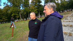 Dwaj mężczyźni stoją na zboczu cmentarza wojennego i obserwują teren porządkowany podczas akcji. W tle widać rzędy odnowionych krzyży oraz osobę filmującą przebieg wydarzenia.