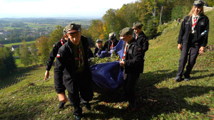Grupa młodych harcerzy niesie duży niebieski brezent wypełniony gałęziami i liśćmi, wchodząc po stromym stoku cmentarza. W tle rozciąga się panorama okolicznych wsi i pól.