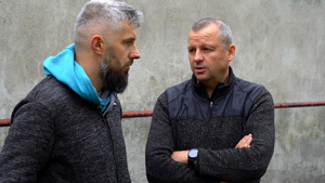 Dwóch mężczyzn w ciemnych, sportowych kurtkach stoi przy jasnym betonowym murze i rozmawia. Po lewej stronie mężczyzna z krótkimi, siwiejącymi włosami i długą siwą brodą, ma turkusowy kaptur bluzy wyciągnięty spod kurtki. Po prawej mężczyzna w ciemnej kurtce z panelami na ramionach, ręce ma skrzyżowane na piersi, słucha rozmówcy.