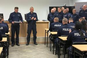 policjanci siedzą w ławkach i słuchają wypowiedzi stojącego przed nimi Komendanta