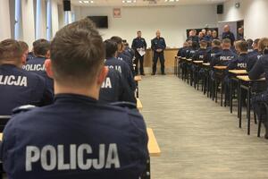 policjanci siedzą w ławkach i słuchają wypowiedzi stojącego przed nimi Komendanta