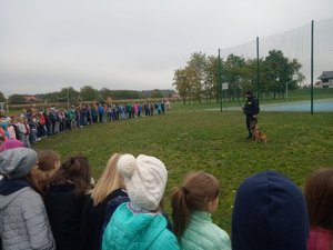 Boisko szkolne. Policjant wraz z psem rozmawia ze zgromadzonymi w półokręgu dziećmi.