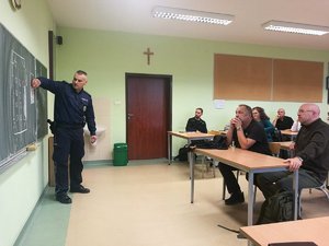 Uczestnicy podczas zajęć w sali szkolnej. Policjant rozrysowuje na tablicy algorytmy postępowania.