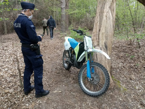 Policjant stoi przy motocyklu opartym o drzewo w lesie.