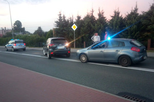 na ulicy stoi skradziona mazda, a obok stoją dwa radiowozy oznakowany po lewej i nieoznakowany po prawej. Na chodniku stoją kryminalni