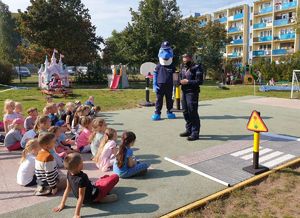 Policjanci prowadzą zajęcia z przedszkolakami na terenie zielonym. Obok stoją elementy miasteczka ruchu drogowego.