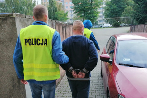 policjant trzymając zatrzymanego skutego kajdankami założonymi na ręce trzymane z tyłu idzie w kierunku samochodu