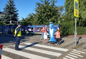 Dwoje dzieci w towarzystwie polfinka przechodzi przez przejście dla pieszych. Przygląda się temu policjant stojący na przejściu dla pieszych.