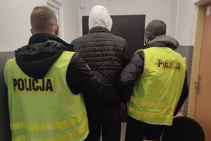 dwaj policjanci stojący tyłem, trzymają zatrzymanego skutego kajdankami