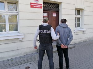 Policjant prowadzi zatrzymanego do prokuratury. W tle budynek prokuratury w Nakle nad Notecią.