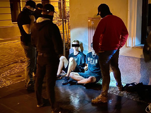 Zatrzymani dwaj mężczyźni w nocy siedzą na płycie starego rynku. Przy nich w półokręgu stoją policjanci po cywilnemu.