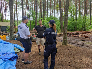 Policjanci rozmawiają z mężczyzną w terenie leśnym. W tle widoczne elementy obozowiska.