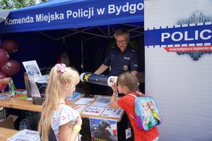 Dwie dziewczynki stoją przy namiocie Komendy miejskiej Policji w Bydgoszczy. Policjant trzyma &quot;magiczną tubę&quot; przed którą patrzy jedna z dziewczynek.