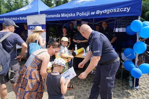 Dzieci i ich opiekunowie przy namiocie profilaktycznym Komendy miejskiej Policji w Bydgoszczy. Policjant rozmawia z dziećmi.