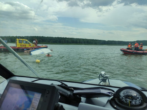 kilka osób trzyma się przewróconej żaglówki. Obok stoi łódź WOPR-u