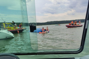 kilka osób trzyma się przewróconej żaglówki. Obok stoi łódź WOPR-u