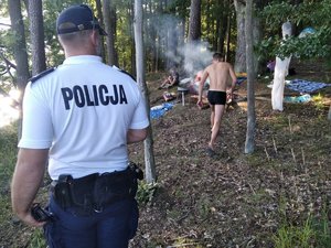 Policjant wchodzi do lasu, gdzie osoby rozpaliły ognisko.  W tle idący mężczyzna i urządzony przez nich biwak.