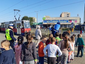 Policjant i Polfinek prowadzą uczniów przez przejścia dla pieszych. Po lewej stronie widoczny nadjeżdżający tramwaj.