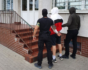 Policjanci prowadzą zatrzymanego do komisariatu. Ujęcie przed schodami prowadzącymi do budynku.