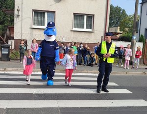 Polfinek z dwojgiem dzieci przechodzi przez pasy pod okiem stojącego policjanta. W tle budynek i grupa dzieci.