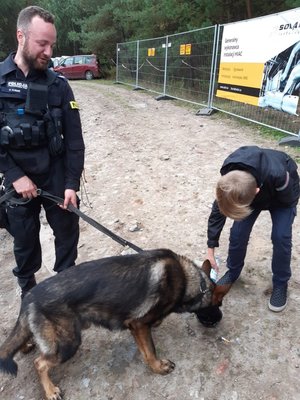 Chłopiec daje wodę do picia psu policyjnemu, którego trzyma na smyczy policjant- przewodnik.