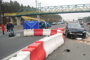 czarny samochód stoi rozbity na drodze. Obok przejeżdżają inne auta. W oddali widać niebieski parawan z napisem Policja