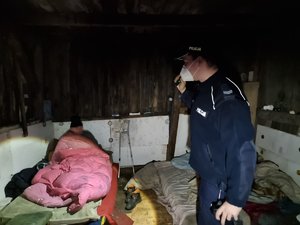 Policjant rozmawia w pustostanie z bezdomnym, który siedzi w łóżku przykryty nakryciami.