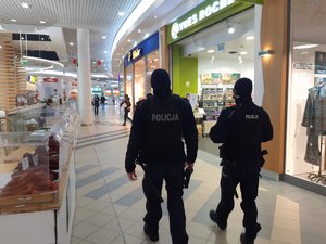 Widok z tyłu na policjantów idących przy stoiskach z wędlinami widocznymi z lewej strony zdjęcia. Po prawej sklepy.