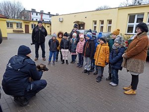 Dzieci i wychowawcy stoją w grupie podczas zajęć na świeżym powietrzu prowadzonych przez policjanta z psem służbowym.