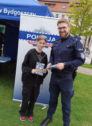 Policjant, a obok niego stoi nastolatka trzymająca w rękach ulotkę.