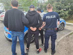 dwaj policjanci trzymają dwóch zatrzymanych skutych kajdankami przed radiowozem