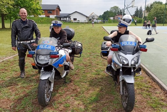 dwaj chłopcy siedzą na dwóch policyjnych motocyklach. Obok stoi policjant