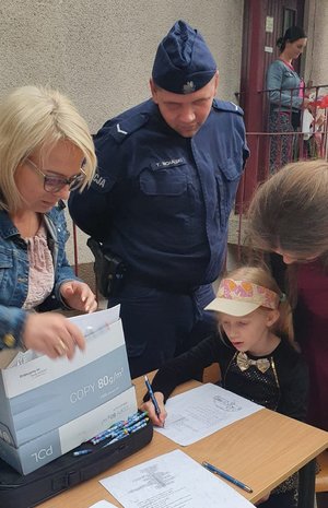 Policjant i kobieta przyglądają się piszącemu przy stoliku dziecku.