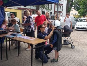 Policjantka zakłada dziecku strój wykorzystywany podczas zabezpieczenia imprez masowych. W tle inne dzieci i dorośli.