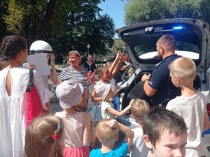 Dzieci i dorośli stoją przy radiowozie od strony bagażnika. Tam też stoi policjant.