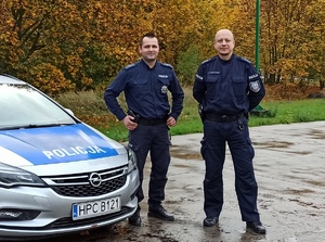 Policjanci stojący obok radiowozu w terenie zielonym.