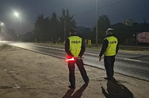 Policjanci stoją przy drodze. Jeden z nich trzyma latarkę do zatrzymywania pojazdów.