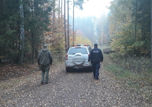 Policjant i leśnik idą w stronę pojazdu służbowego zaparkowanego w lesie.