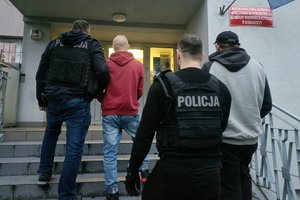 dwaj nieumundurowani policjanci w kamizelkach taktycznych z napisem Policja prowadzą po jednym zatrzymanym skutych kajdankami